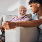 Stock image of older male adult sitting next to younger male representing a caregiver relationship.